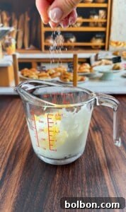 A hand sprinkles salt into a glass measuring cup containing a white liquid, likely cream or milk, on a wooden table with a blurred background of baked goods and what could be ingredients for Pizza Crouton Salad.