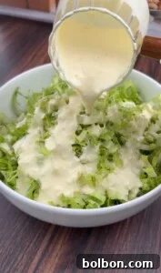 A creamy dressing is being poured from a glass pitcher onto a bowl of shredded lettuce, creating a delicious Pizza Crouton Salad on a wooden table.