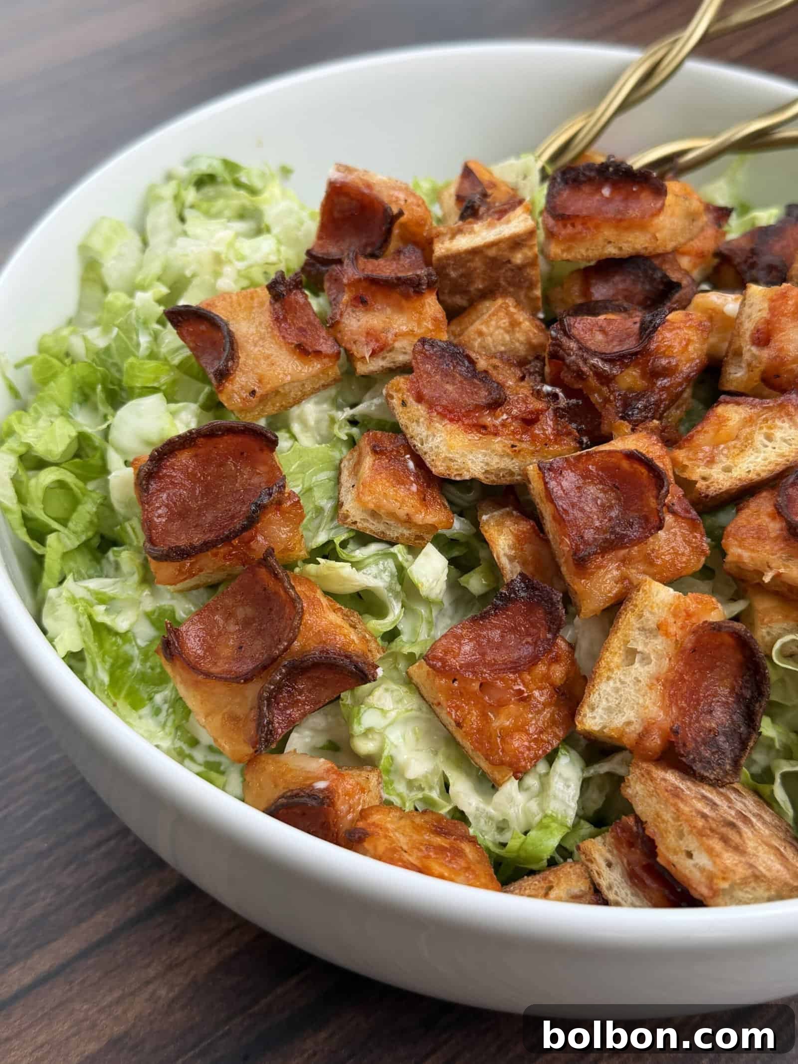 A close-up of a Pizza Crouton Salad in a white bowl, topped with crispy, browned croutons and diced vegetables on shredded lettuce. Two gold utensils rest inside the bowl on a wooden surface.