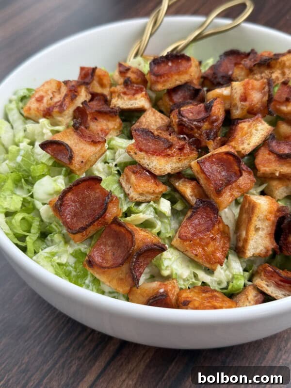 A close-up of a Pizza Crouton Salad in a white bowl, topped with crispy, roasted bread cubes and thin slices of pepperoni. The green salad underneath appears to be chopped lettuce. Two gold-colored utensils rest in the bowl.