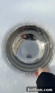 A hand holds a metal bowl partially embedded in snow, with a reflection of a person and buildings on its shiny surface. The scene is bright, set against a soft white snowy background—perfect for making viral snow ice cream.