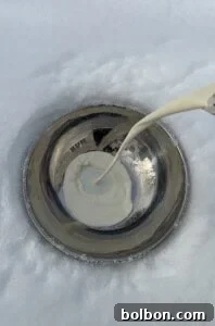 A stream of cream is being poured into a metal bowl placed on snow, with the cream beginning to pool in the center—perfect for making Viral Snow Ice Cream.
