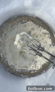 A close-up of an electric mixer with beaters covered in Viral Snow Ice Cream, held above a metal mixing bowl filled with the same creamy mixture.