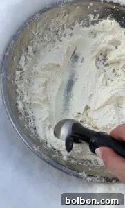 A hand holding an ice cream scoop scrapes Viral Snow Ice Cream from a metal mixing bowl, with snow visible around the bowl's base.