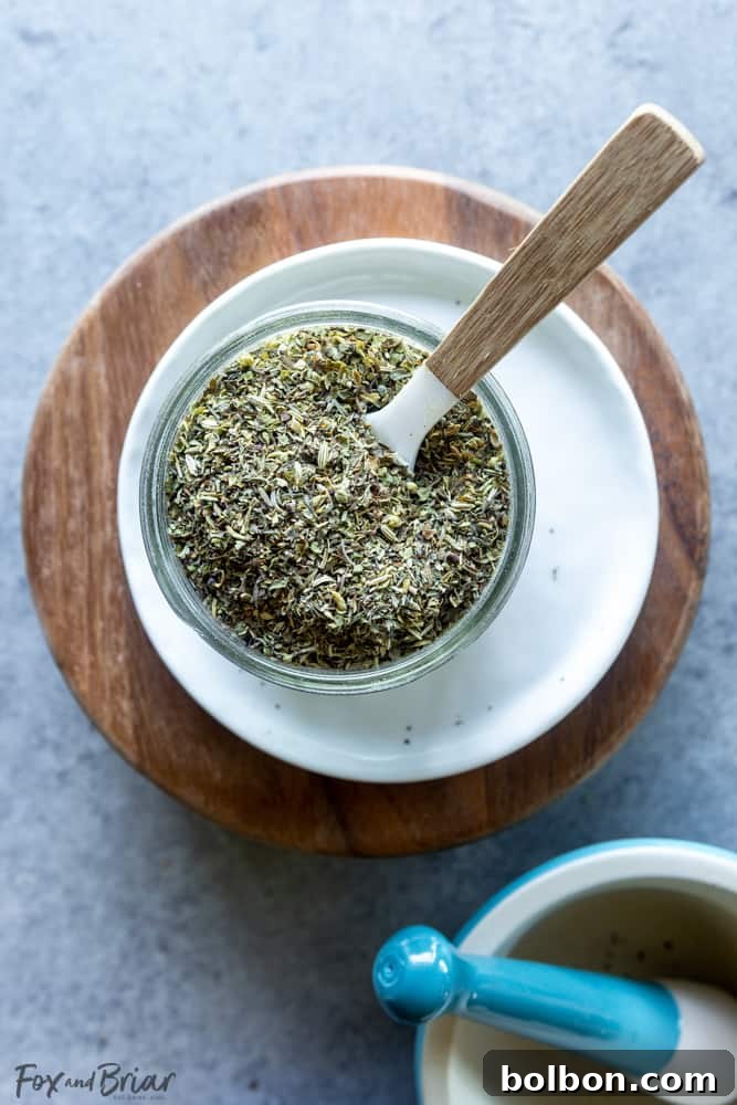 A hand holding a wooden spoon filled with homemade Italian seasoning over a bowl of dried herbs.
