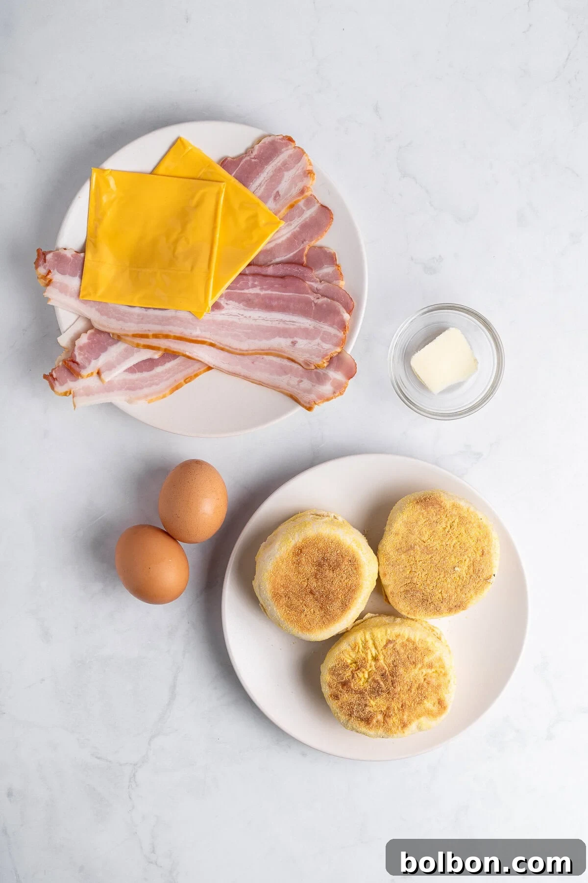 A selection of raw ingredients for homemade Egg McMuffins laid out on a marble surface, including uncooked bacon slices, slices of cheddar cheese, two fresh eggs, butter, and two halved English muffins.