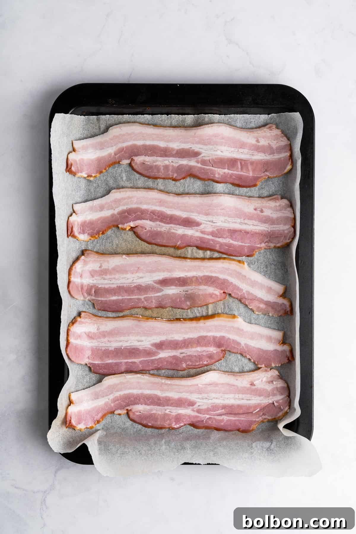 Uncooked bacon strips, neatly arranged in a single layer on a parchment-lined baking tray, ready to be placed in the oven for cooking. The tray sits on a light marble surface.