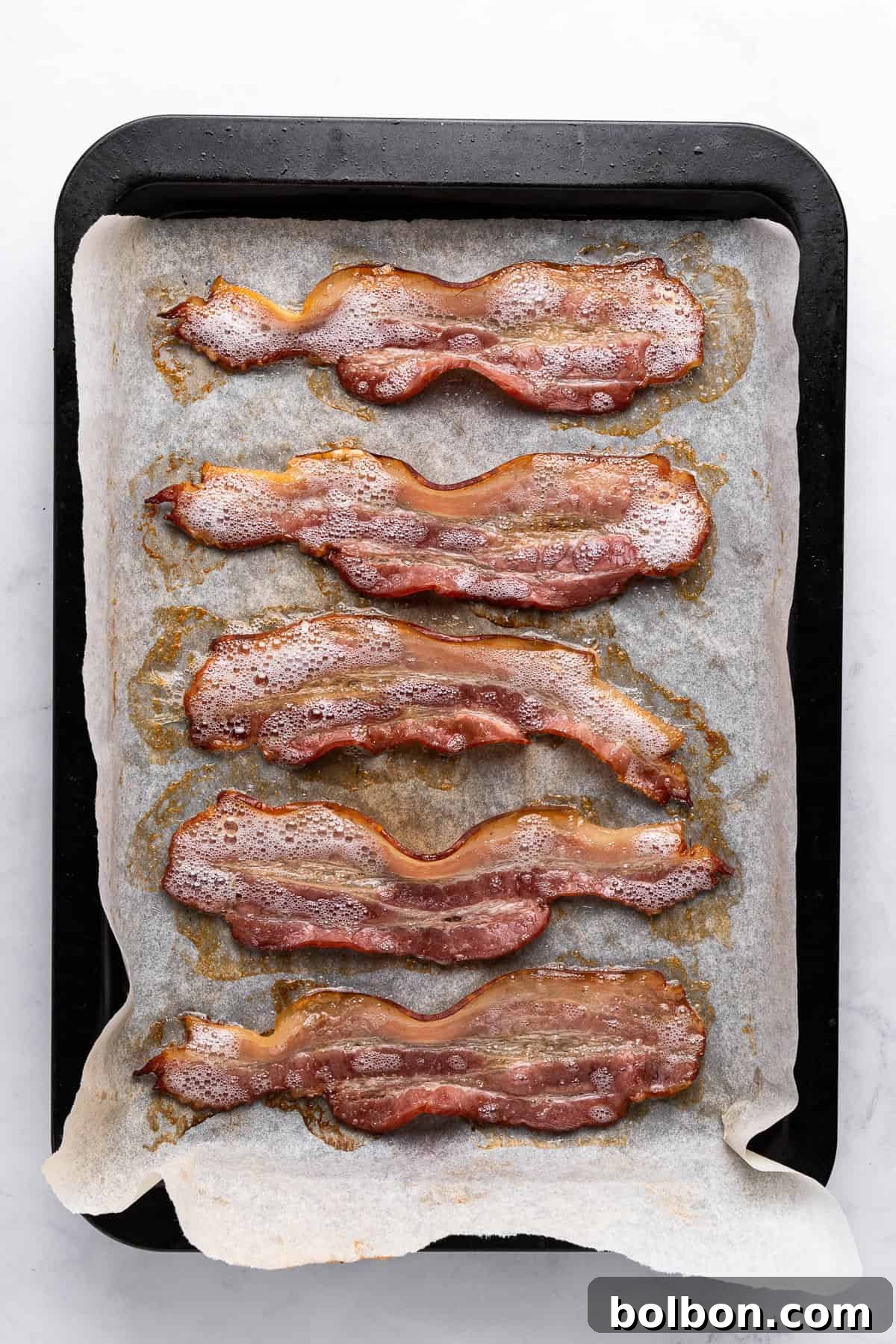 Five slices of perfectly cooked, crispy bacon resting on a baking tray lined with parchment paper, having achieved an even golden-brown color. The tray is positioned on a light countertop.
