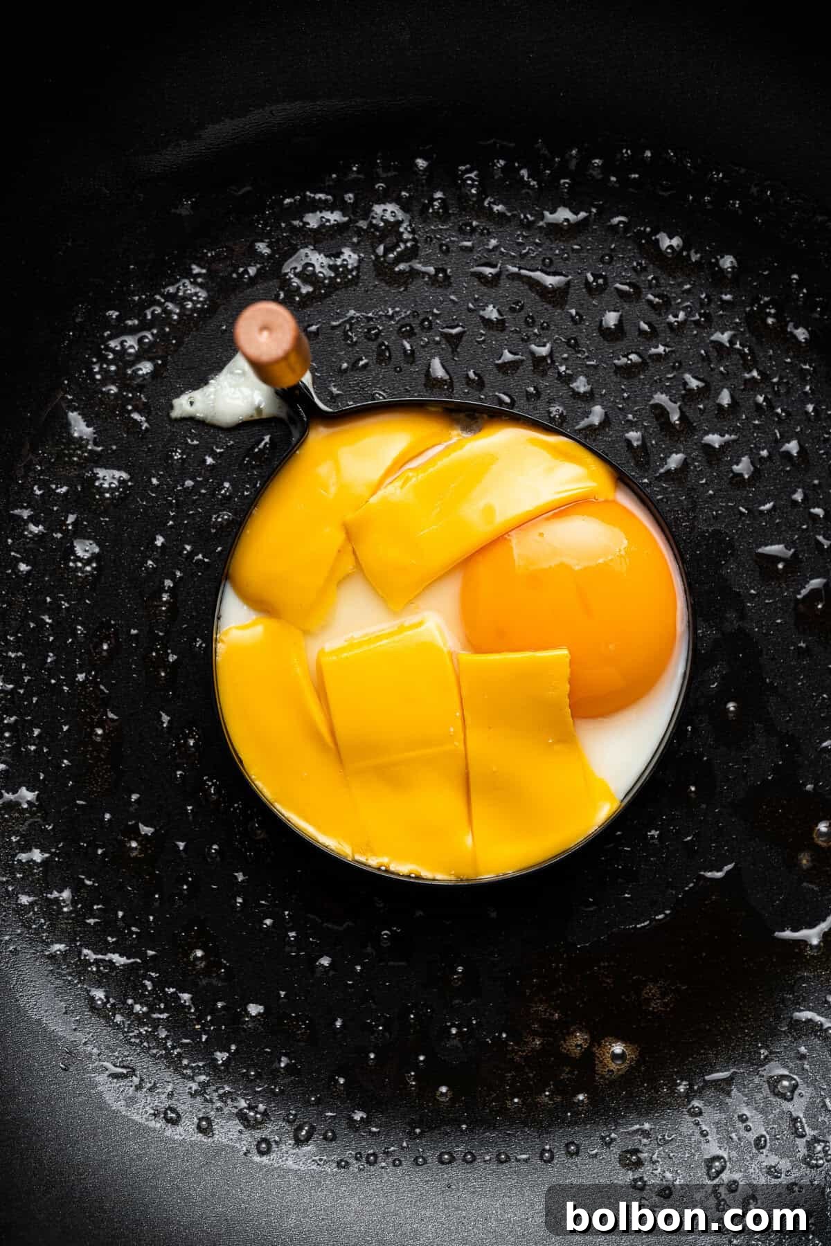 A partially cooked egg with a golden yolk and neatly trimmed egg whites in a non-stick frying pan, glistening with oil droplets. A cracked eggshell sits beside it, indicating fresh preparation.
