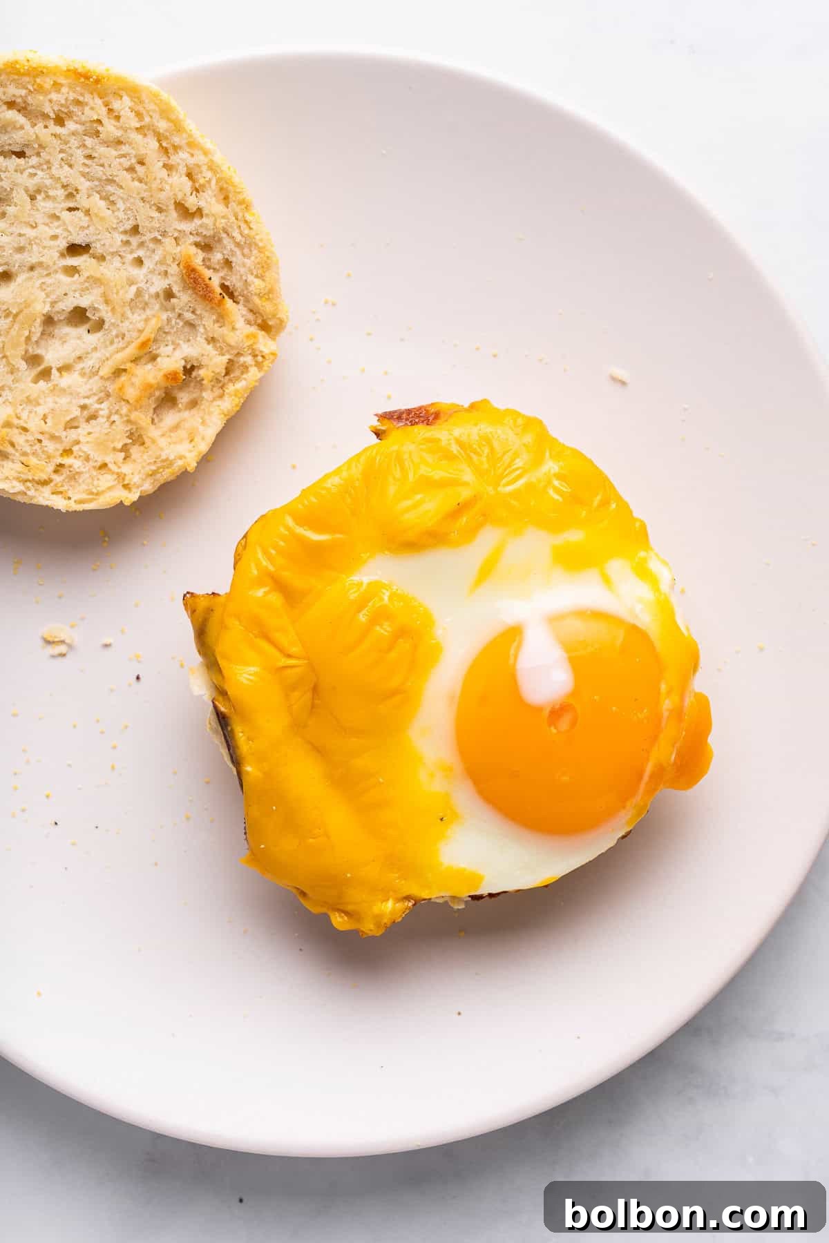 A single toasted English muffin half, topped with a perfectly fried egg and melted cheese, with a hint of runny yolk, served on a white plate alongside a slice of plain bread. The background is simple and bright.