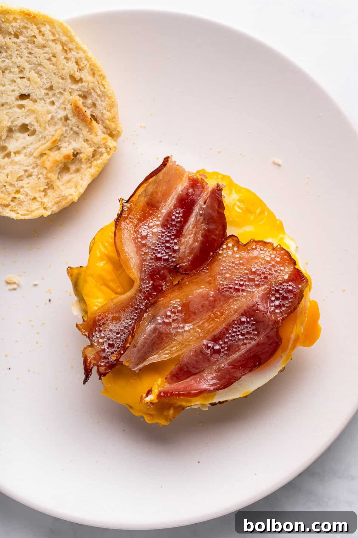 The final step of assembling a homemade Egg McMuffin: crispy bacon is layered onto a fried egg with melted cheese, all resting on a toasted English muffin. The sandwich is presented on a white plate with a side of bread.