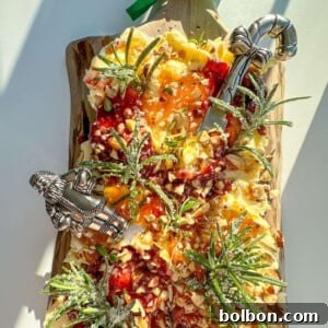 A festive Holiday Butter Board topped with nuts, fruit preserves, herbs, and sugared rosemary sprigs, arranged on a wooden board with two decorative cheese knives and a green ribbon bow.