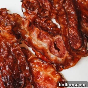 Crispy, cooked strips of bacon arranged on a white surface showcase how to cook bacon in the oven for a rich brown color and glossy, caramelized texture with slightly curled edges.