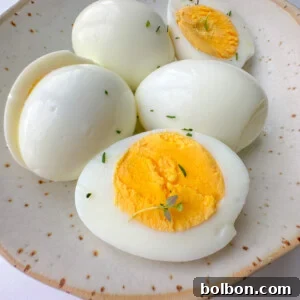 A ceramic plate holds four Easy Peel Hard Boiled Eggs, two sliced in half to reveal their bright yellow yolks and garnished with small green herb sprigs.