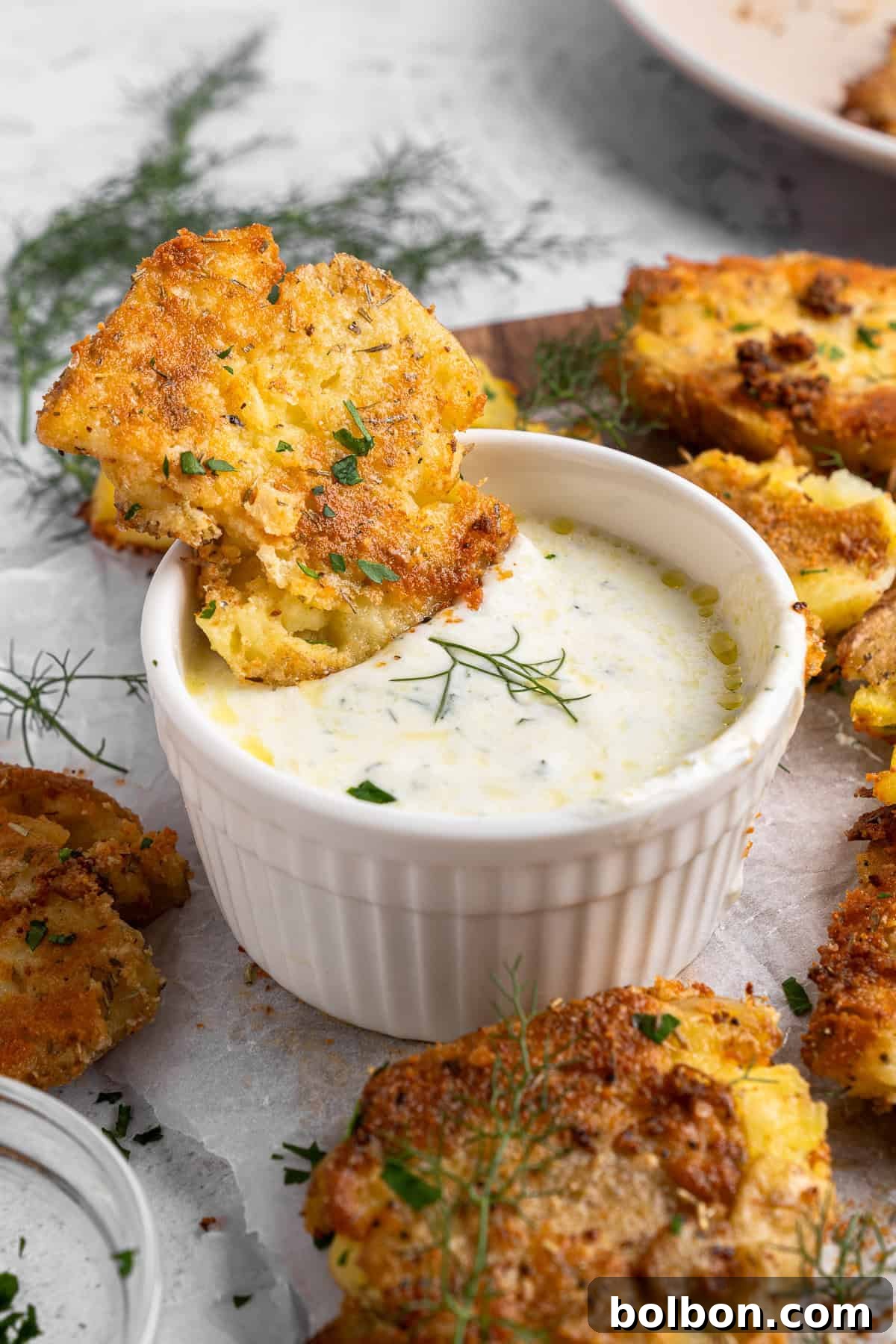 A piece of fried corn fritter is dipped into a small white ramekin filled with creamy herb sauce. Additional fritters are scattered around on a light surface, garnished with fresh dill leaves.