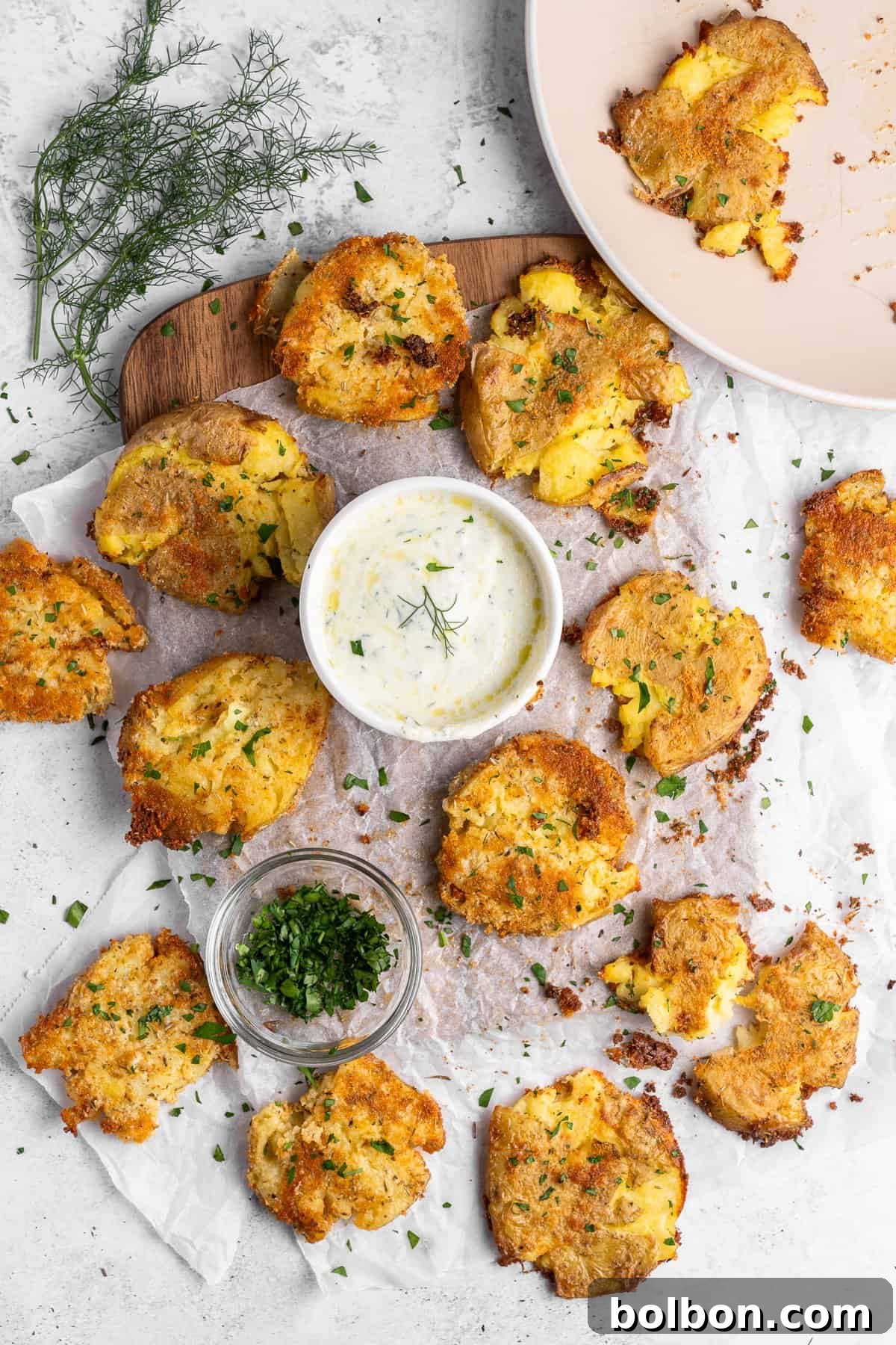 Golden brown smashed potatoes sprinkled with herbs are arranged on parchment paper. A small white bowl of creamy dip garnished with dill is centered on the board. Extra chopped herbs are in a glass jar nearby. A plate with some eaten potatoes is on the side.