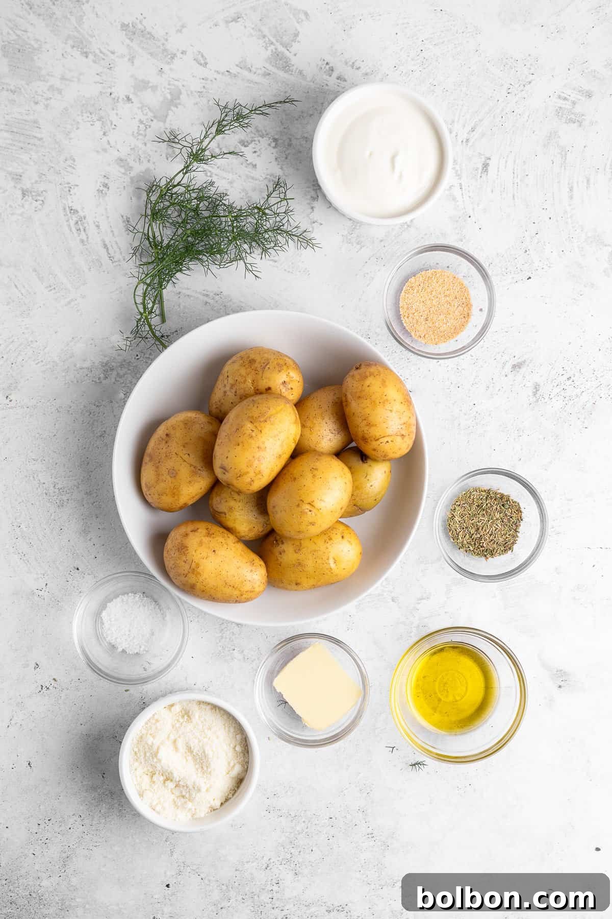 A bowl of potatoes is surrounded by small bowls containing various ingredients: sour cream, dill, garlic powder, dried herbs, olive oil, grated cheese, butter, and salt on a light textured surface.