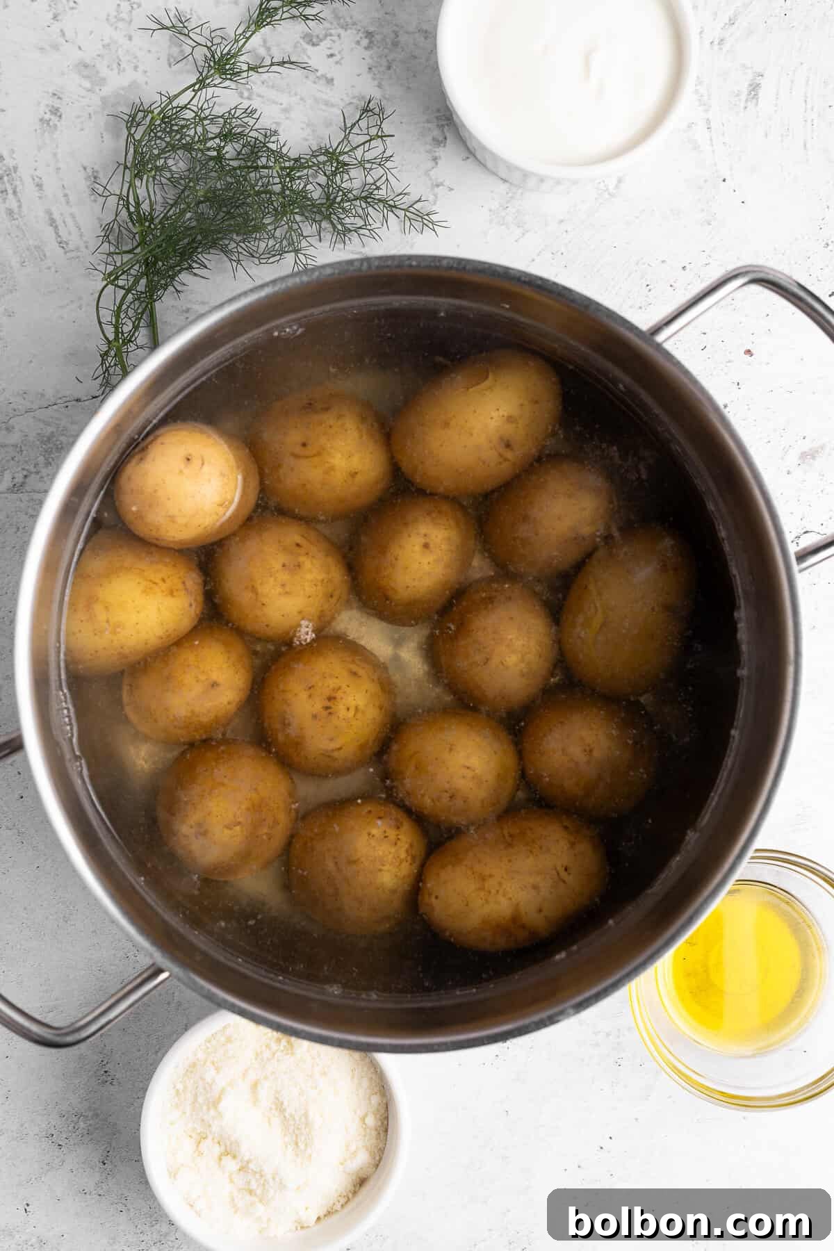A pot filled with boiling potatoes is centered in the image. Surrounding the pot are small bowls containing butter, flour, and sour cream. Fresh dill is placed nearby on the textured white surface.