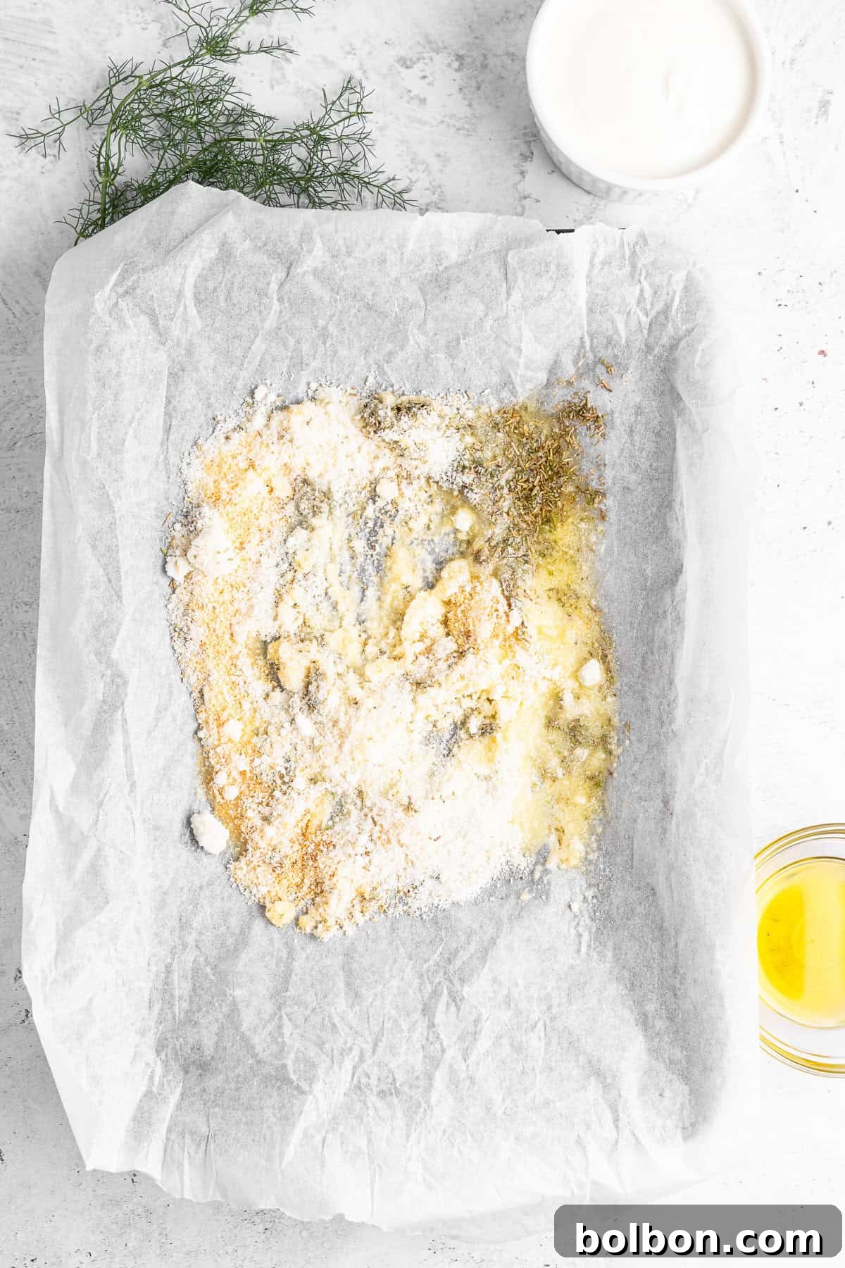 A baking tray lined with parchment paper contains mixed spices, flour, and yogurt, creating a marbled texture. Fresh dill is placed nearby, with a small bowl of yogurt and a glass bowl of olive oil on the side.