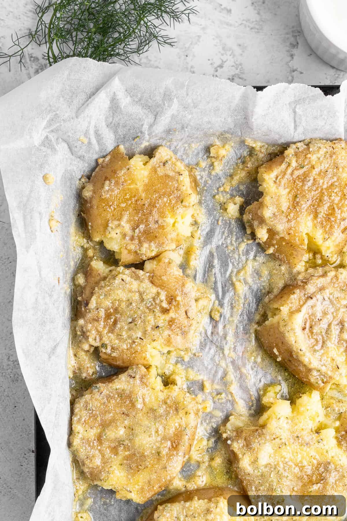 Crushed potatoes on a parchment-lined baking tray, seasoned with herbs and golden-brown baked, with a sprig of fresh dill in the background.