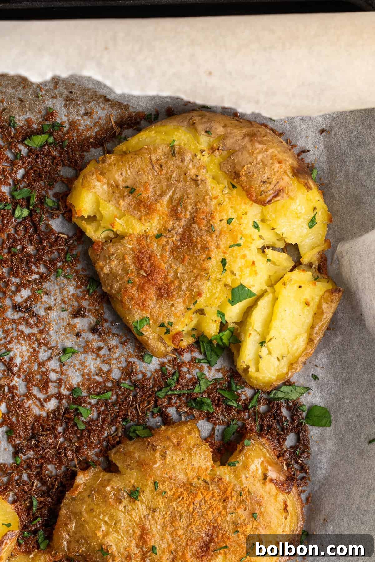 A close-up of crispy potatoes garnished with chopped parsley. The potatoes are golden brown with a textured surface, resting on parchment paper, with bits of seasoning visible around them.