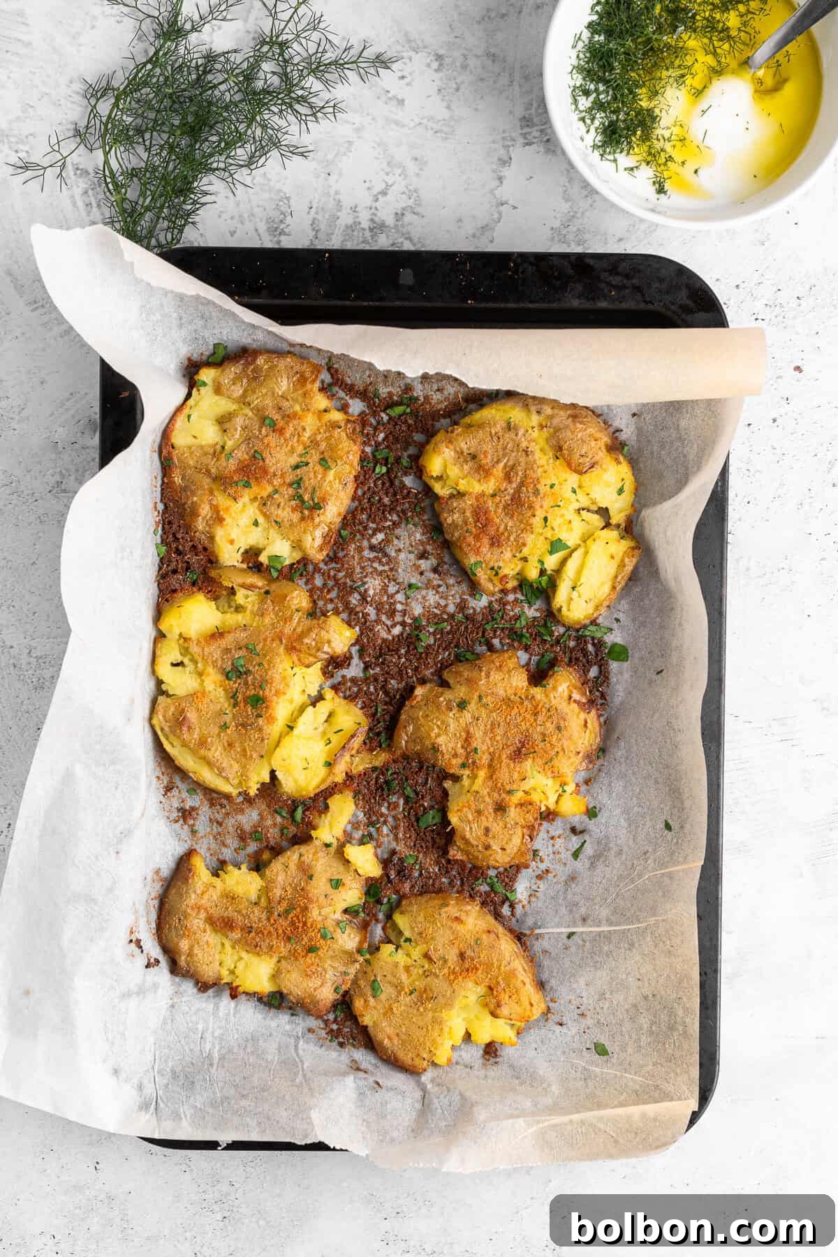 Baked smashed potatoes on a parchment-lined tray, garnished with herbs. A small bowl with olive oil and herbs is in the background. The setting is on a light gray surface with a sprig of fresh herbs nearby.