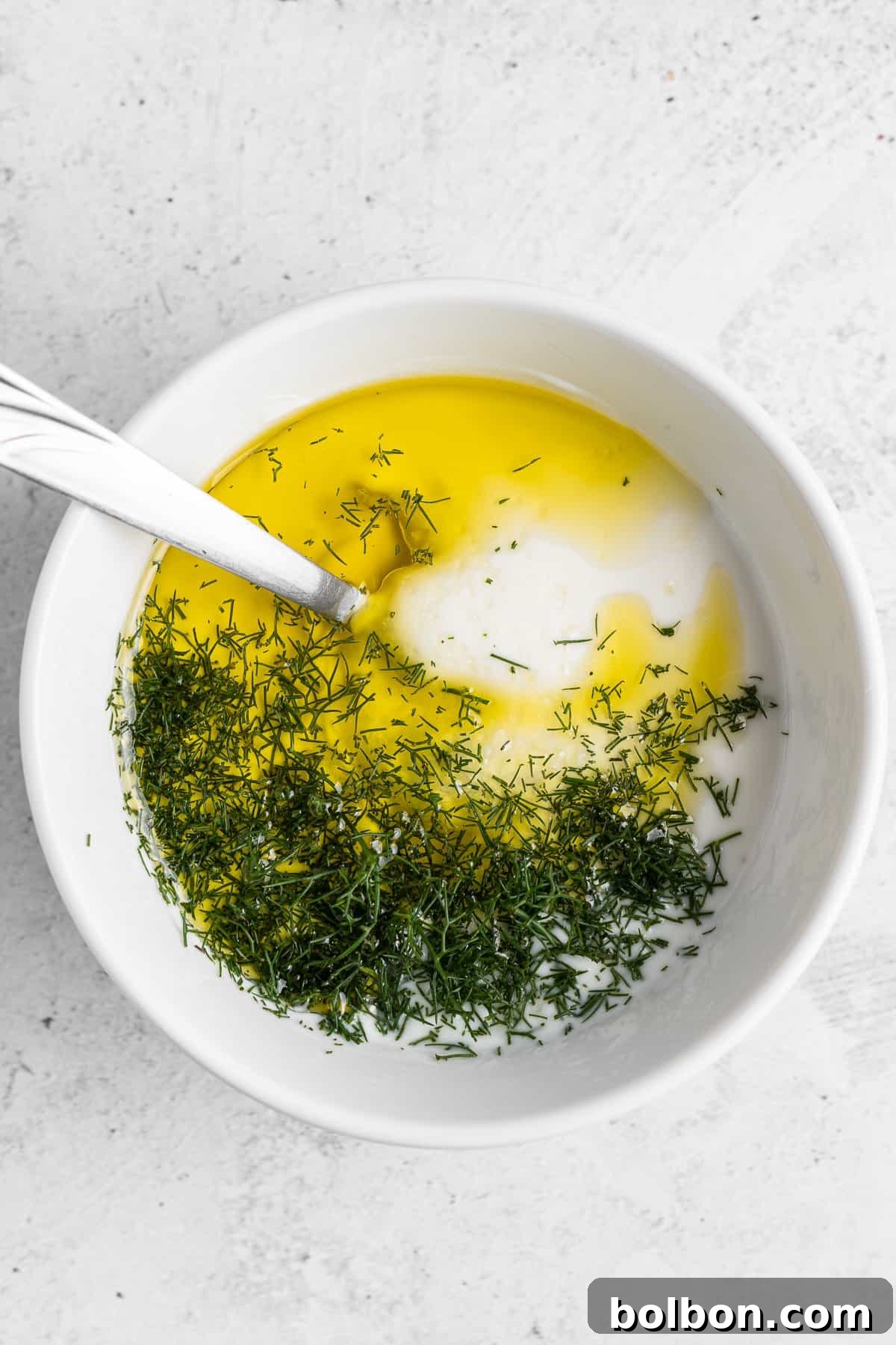 A white bowl containing yogurt, olive oil, and dill, with a spoon resting inside. The ingredients are partially mixed, creating a vibrant contrast of colors and textures. The background is a light, textured surface.