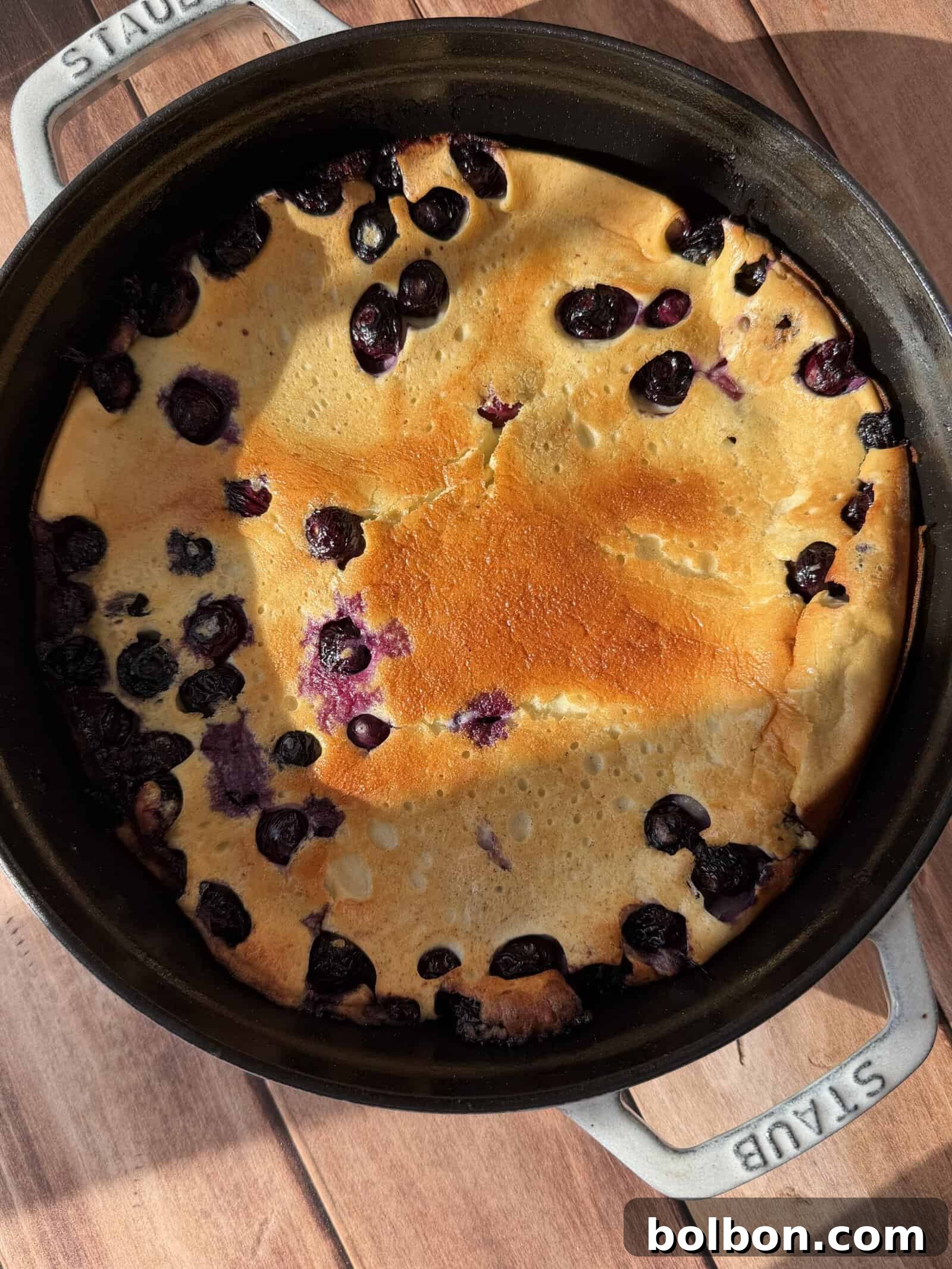 Protein-Packed Dutch Pancake 2 A High Protein Dutch Baby, golden-brown and baked with blueberries, is presented in a round black Staub baking dish on a wooden surface, sunlight casting soft shadows across the scene.