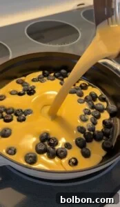 Protein-Packed Dutch Pancake 7 A creamy high protein Dutch baby batter is being poured into a pan full of fresh blueberries, preparing a dessert on a stovetop.