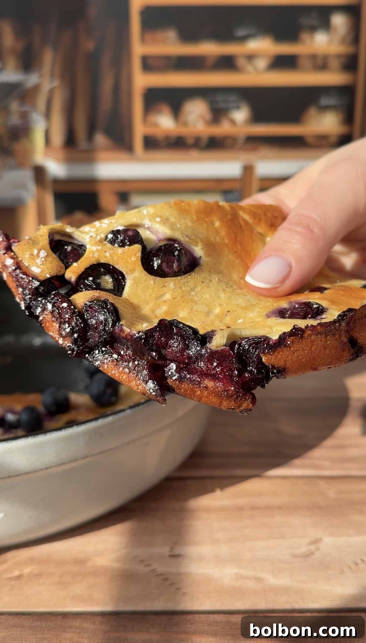 Protein-Packed Dutch Pancake 3 A hand holds a thick, golden-brown slice of High Protein Dutch Baby pancake, with juicy blueberries visible throughout. A ceramic dish with more pancake is in the background on a wooden table.