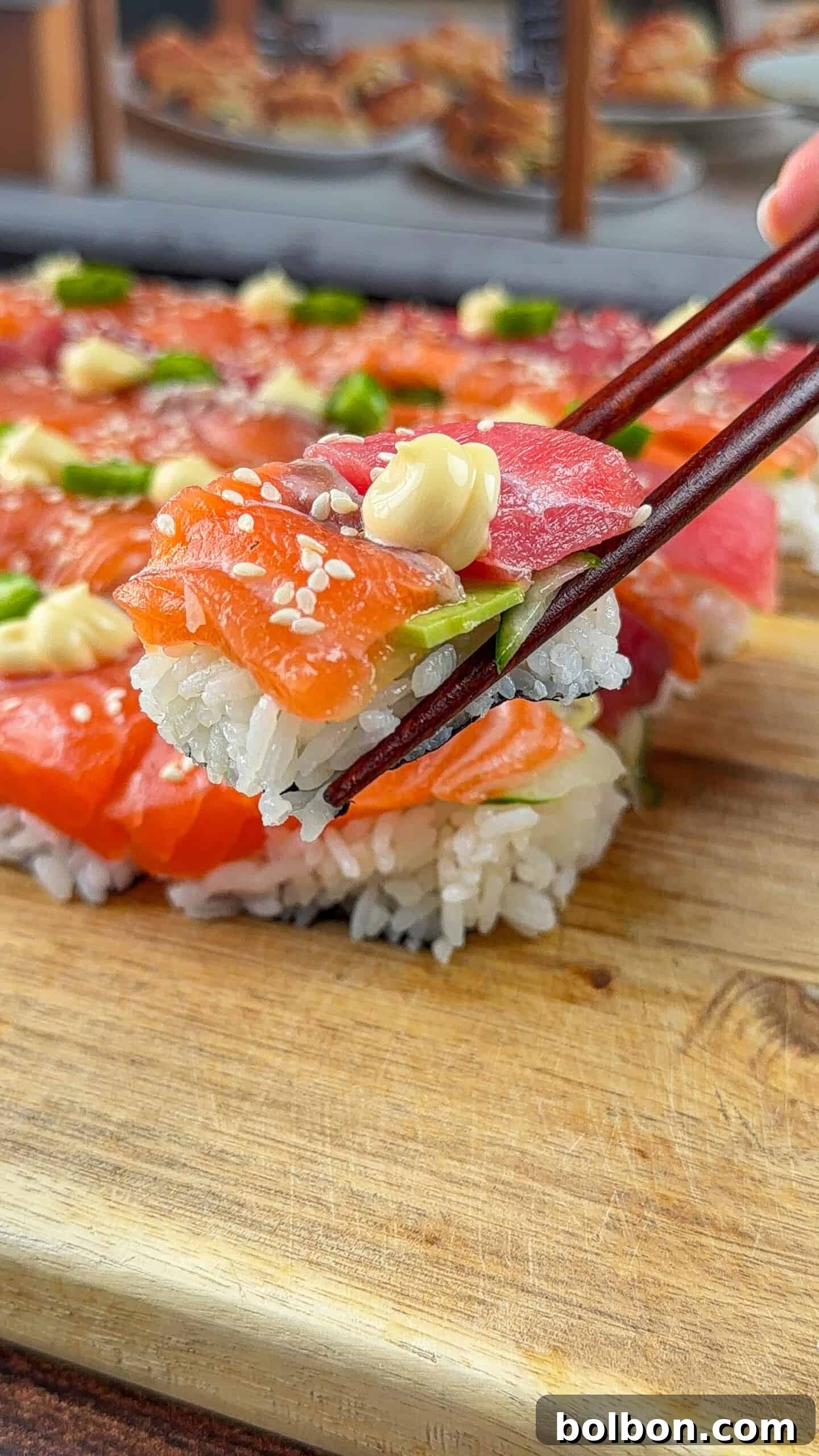 The Viral One Pot Sushi Cake Phenomenon 2 A close-up of chopsticks holding a piece of sushi with rice, fresh salmon, tuna, green onions, sesame seeds, and a dollop of mayonnaise, above a wooden board with more sushi in the background.