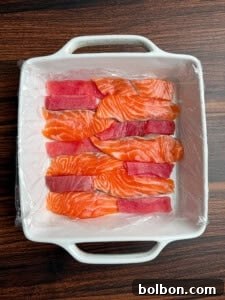 A white square dish lined with plastic wrap holds alternating rows of sliced raw salmon and tuna, displayed neatly on a dark wooden surface.