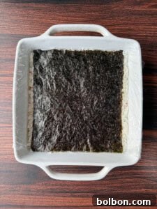 A square white dish with handles contains a layer of white rice topped with a sheet of nori seaweed, partially covered with plastic wrap, on a dark wooden surface.