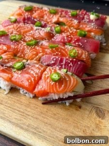 Rectangular pieces of sushi with salmon and tuna slices on rice, topped with green chili slices and sesame seeds, arranged on a wooden board. A pair of chopsticks holds one piece.