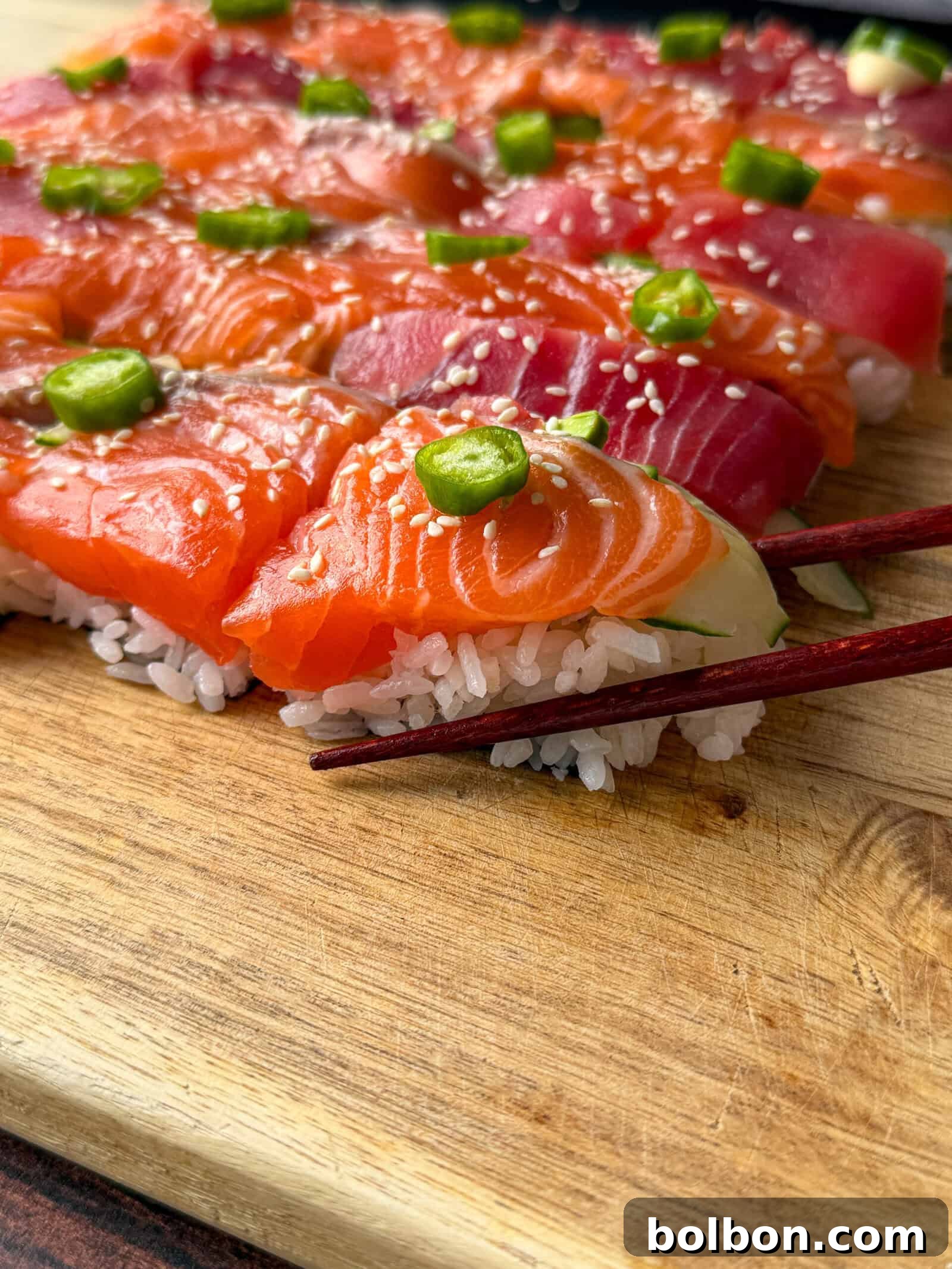 The Viral One Pot Sushi Cake Phenomenon 3 Close-up of assorted sushi topped with fresh slices of salmon and tuna, green wasabi dollops, and sesame seeds, arranged on a wooden board with a pair of chopsticks picking up a piece.