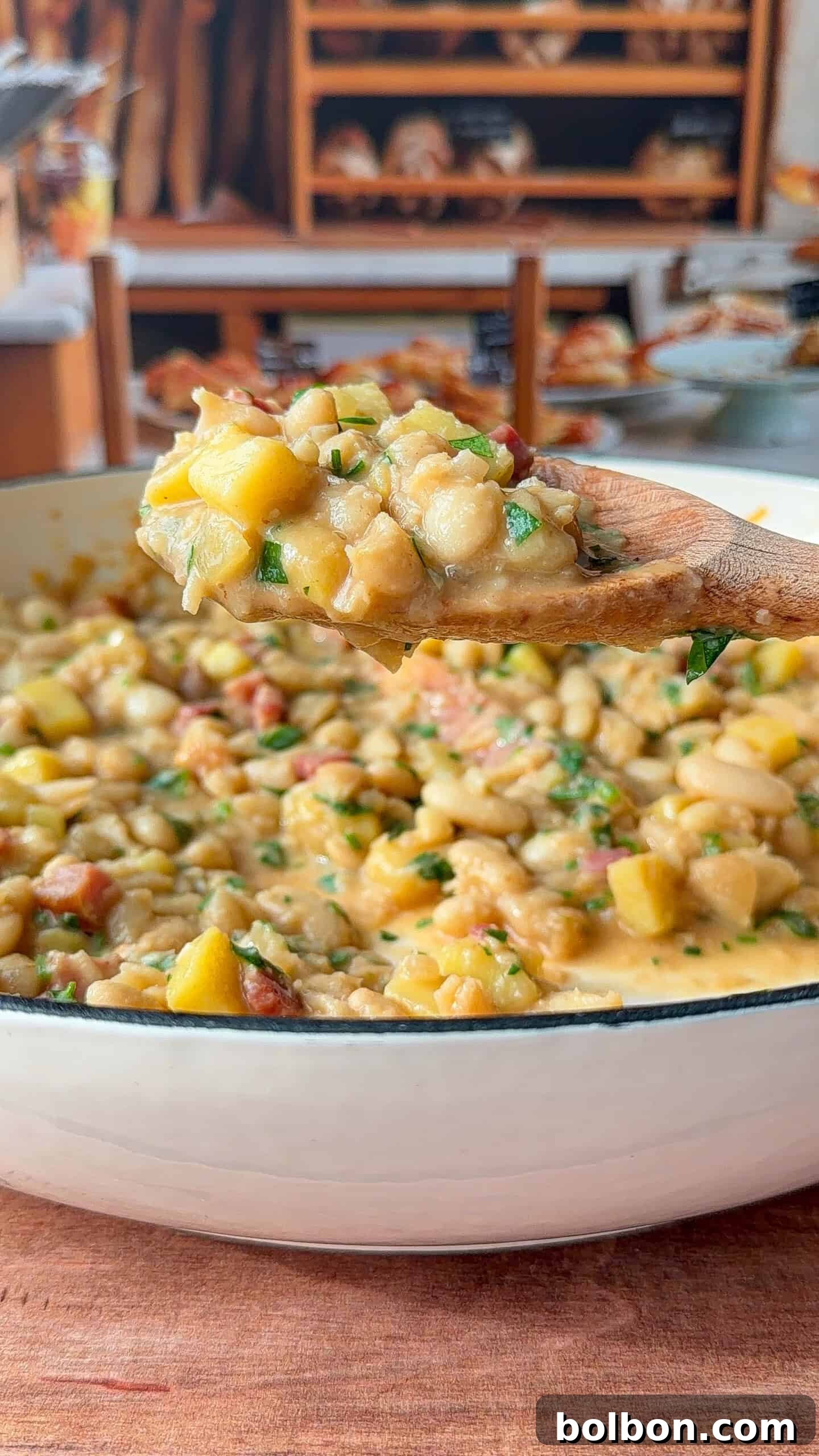 A wooden spoon holds a scoop of creamy bean and potato stew over a white pot, with visible chunks of yellow squash, beans, and herbs. In the blurred background, shelves display various breads and baked goods.