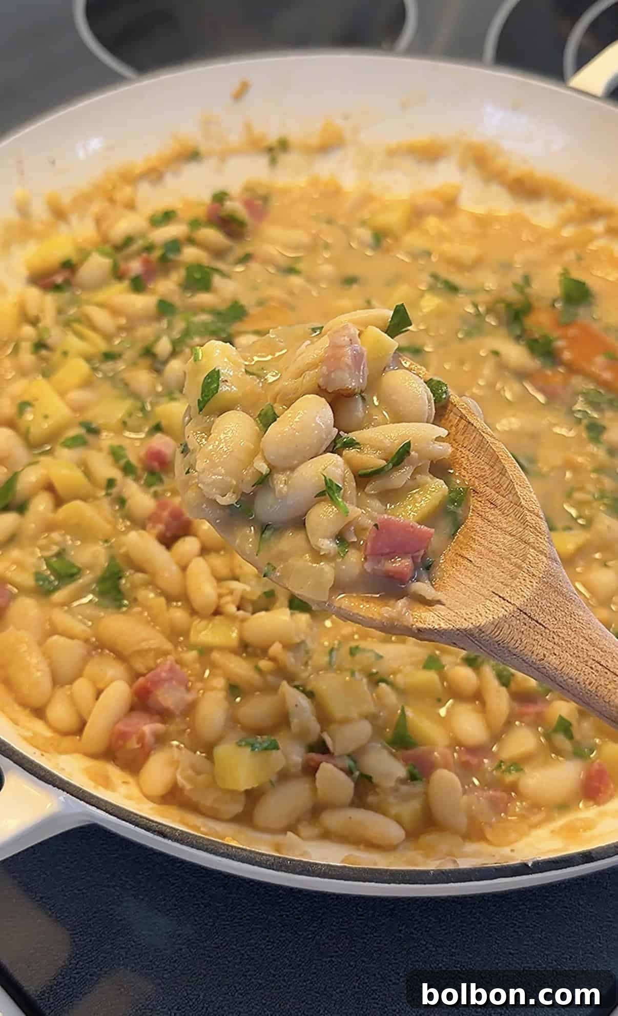 A wooden spoon holds up a serving of Creamy Bean and Potato Stew with chopped herbs and diced ham over a pot filled with the same creamy mixture.