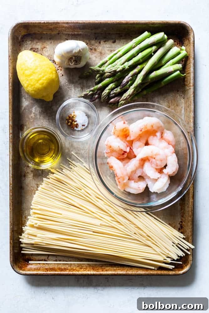 Fresh ingredients for Shrimp and Asparagus Pasta, including green asparagus spears, a bright yellow lemon, cloves of garlic, a bottle of olive oil, salt, red pepper flakes, and raw pink shrimp, beside uncooked pasta.