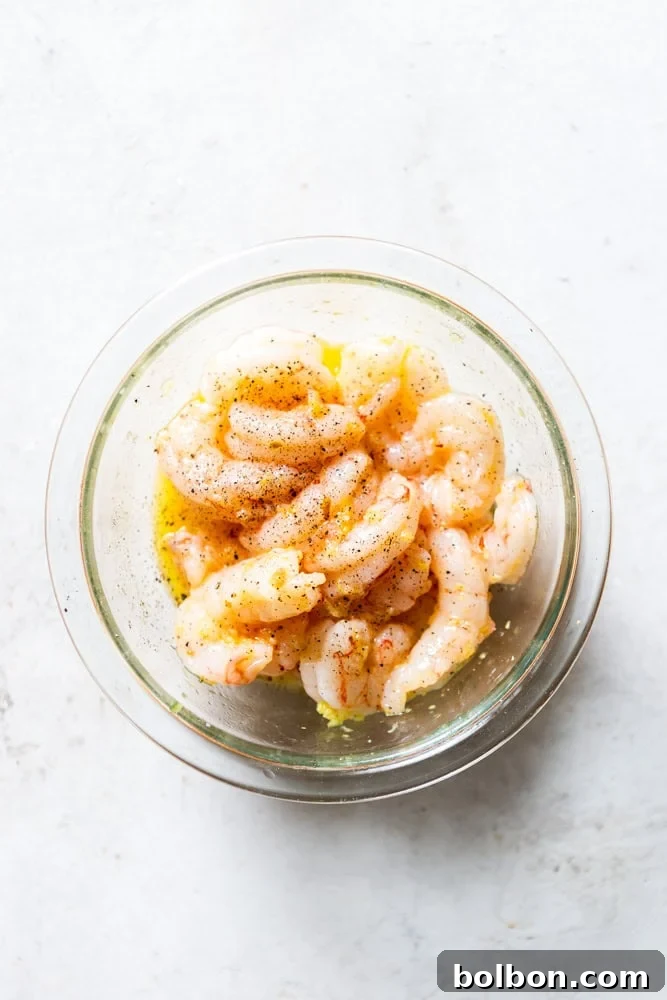 Raw shrimp tossed with olive oil, minced garlic, and lemon zest in a white bowl, ready for roasting.