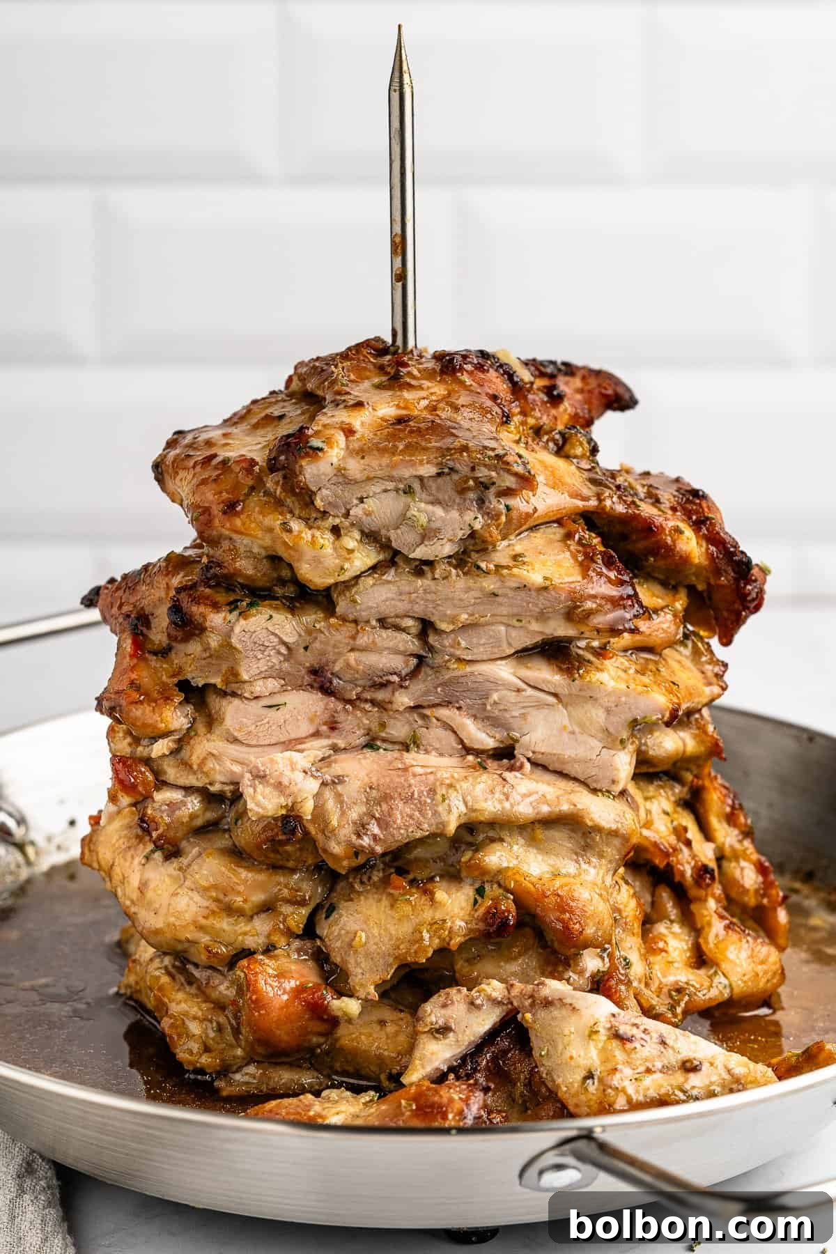 A vertical stack of grilled, seasoned meat slices on a skewer, resembling shawarma or gyro, sits in a metal pan. The high protein gyro is golden brown and juicy, with a crispy outer layer, set against a white tiled background.