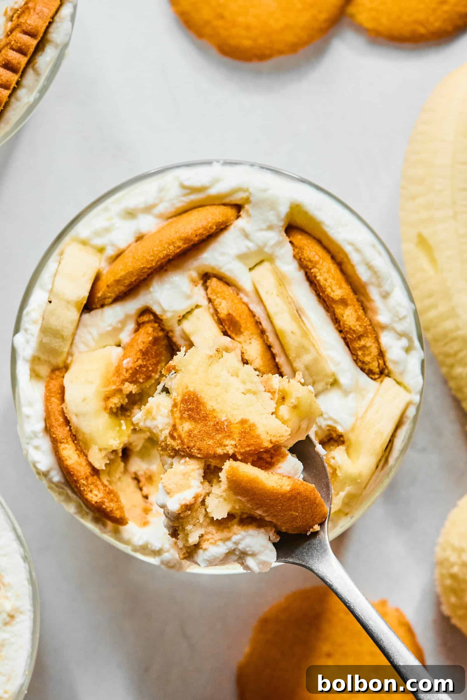 A close-up of banana pudding in a glass dish, topped with vanilla wafers and banana slices, with a spoon scooping out a portion. Vanilla wafers and a banana are visible in the background.