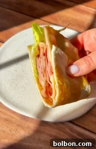 A hand holding a folded wrap filled with healthy mortadella, melted cheese, and crisp lettuce, presented on a white plate on a wooden table, bathed in warm sunlight.