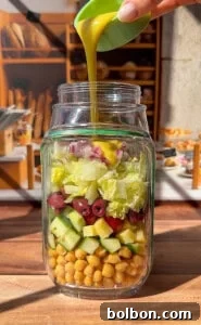 A hand carefully pours golden salad dressing into a clear glass jar filled with perfectly layered chickpeas, cucumbers, olives, lettuce, and finely diced red onions. The jar rests on a warm wooden table in a sunlit kitchen, ready for the next step of meal prep.