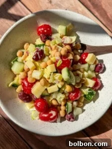 A vibrant, mixed Greek salad served in a white speckled bowl on a natural wooden surface. The salad contains fresh cherry tomatoes, chickpeas, kidney beans, chopped cucumber, crisp lettuce, and small cubes of cheese, showcasing a hearty and healthy meal.