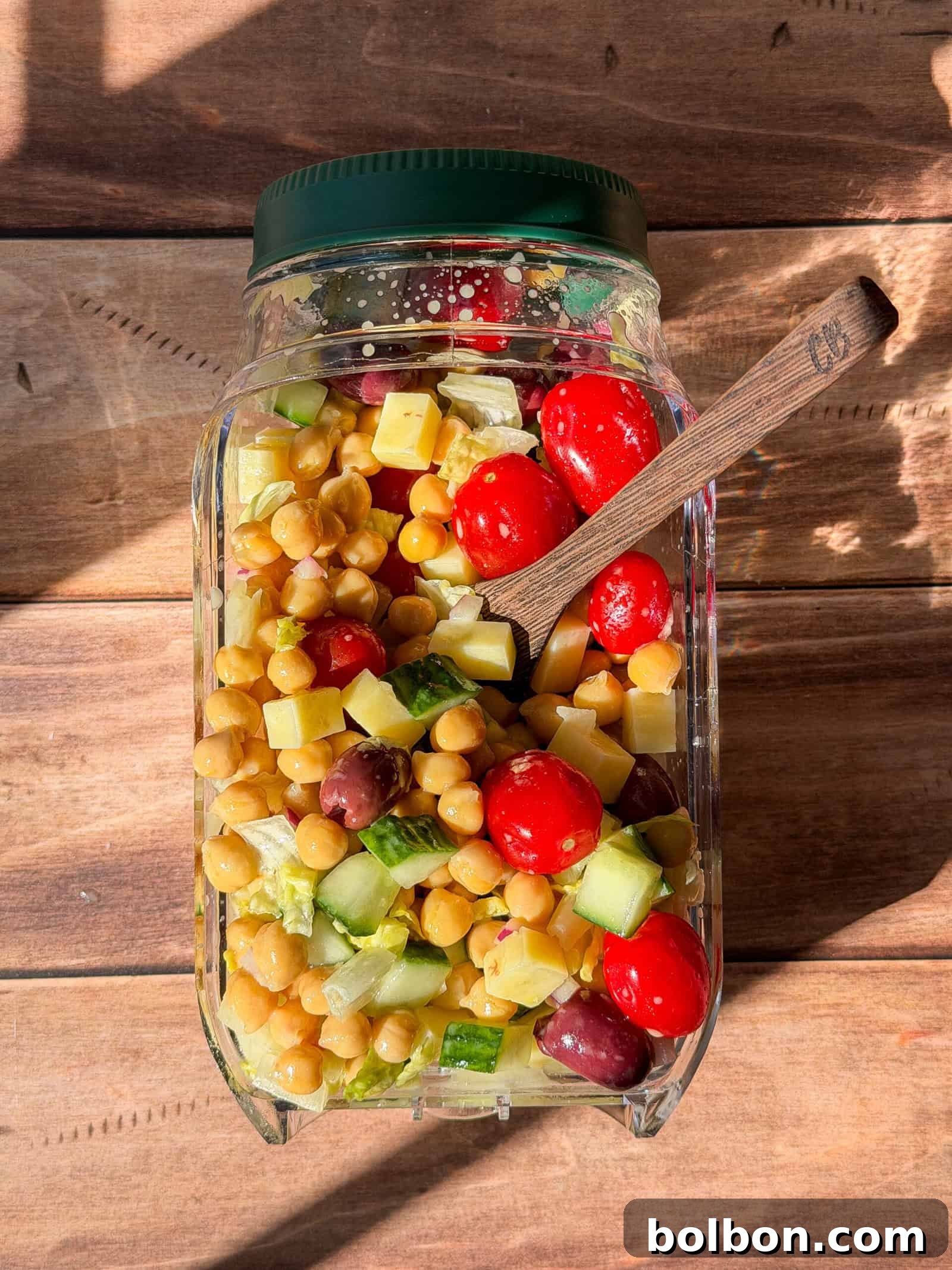 A neatly layered Greek salad in a clear glass jar, featuring chickpeas, cucumbers, cheese cubes, grape tomatoes, and olives, topped with fresh lettuce. The jar is placed on a wooden surface in bright sunlight, with a wooden spoon inside, indicating readiness to eat.