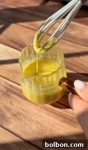 A hand holds a measuring cup of vibrant yellow dressing for a Greek Salad Jar, while a whisk is lifted, allowing the sauce to drip back into the cup on a sunlit wooden surface, emphasizing freshness.