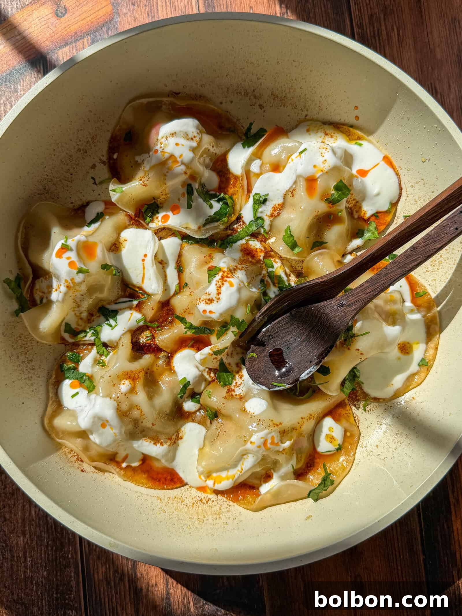A bowl of Viral Blanket Dumplings topped with creamy white sauce, red oil, and chopped herbs sits on a wooden surface. A wooden spoon rests in the bowl under bright natural light.