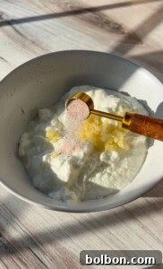 A bowl of white yogurt with finely chopped garlic. A golden measuring spoon holds pink salt, ready to be added to the mixture. The bowl sits on a light wooden surface with soft shadows.