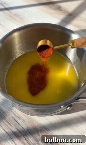 A tablespoon of red spice is being poured into a saucepan containing melted butter, set on a light-colored wooden surface. The sunlight casts gentle shadows across the scene.