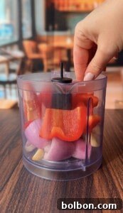 A hand holds the lid of a clear plastic food chopper filled with chopped red bell pepper, red onion, and garlic cloves on a wooden table, with a blurred indoor background.